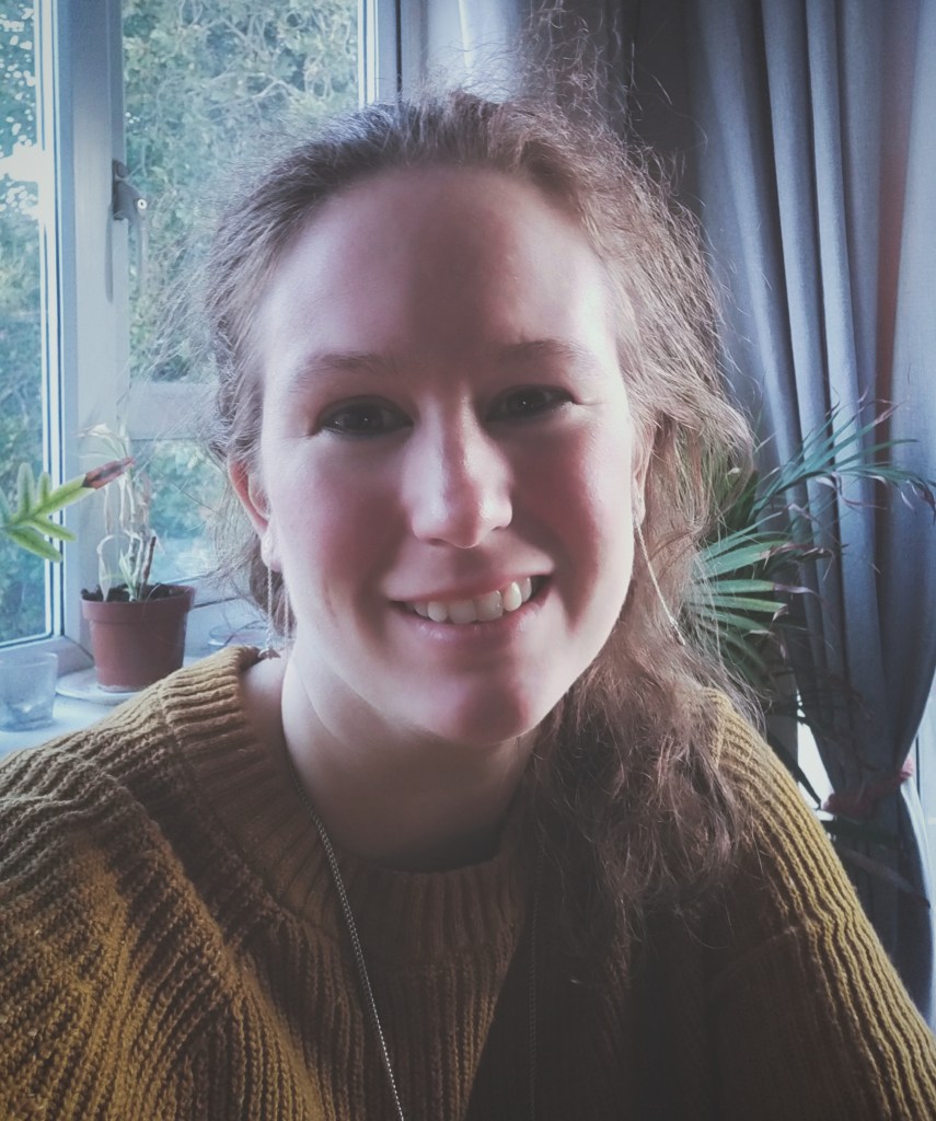 Lucy, a freelance writer, smiling in front of a window. She dark red hair and a mustard yellow jumper
